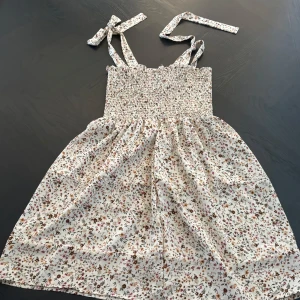 Blommig sommarklänning med smock och knytband - Supersöt blommig klänning med knytbara axelband. Ljus botten med små blommor i olika färger. Perfekt för varma dagar och ger en romantisk känsla.