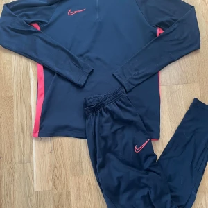 Nike Dri-Fit Academy tracksuit | Tröja + byxor i svart & rött - Komplett Nike Dri-Fit Academy tracksuit med jacka och byxor. Svart med röda detaljer. Både jackan och byxorna ingår. Snygg sportig passform, perfekt till träning eller vardag. Storlek: S (passar dig som är ca 170–180 cm). Använd men i bra skick. Inga fläckar eller hål. Skicka gärna meddelande om du vill ha fler bilder!