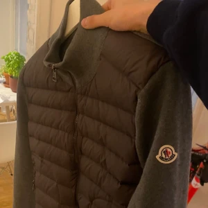 Grå Moncler Cardigan - Säljer en snygg grå cardigan från Moncler med quiltad front och ärmar i stickad ull. Jackan har dragkedja framtill, hög krage och två fickor med dragkedja, ingen luva. Klassisk Moncler-logga på ärmen med NFC och QR kod som skannar, kvitto finns inte då den är köpt second hand men äkthet garanteras:) haft i ungefär ett år så inte ny men 9/10 kvalite, inga defekter eller liknande, storlek XS men passar S också. Säljer då jag växt ur den. Skriv om ni har frågor eller liknande!