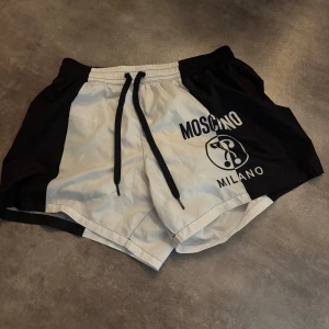 Svarta och vita shorts från Moschino - Snygga shorts från Moschino i svart och vitt med tryckt logga och text på ena benet. De har snörning i midjan och sportig design, perfekta för en trendig look. 