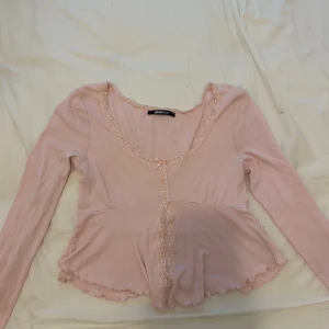 Ljusrosa långärmad topp från Gina Tricot - Söt ljusrosa långärmad topp från Gina Tricot med spetsdetaljer längs ringningen och framtill. Toppen har en liten rosett framtill och volangkant nedtill, vilket ger en romantisk känsla.