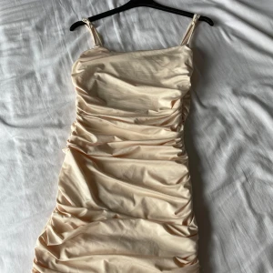 Champagnefärgad omlottklänning med rynkade detaljer - Säljer en snygg champagnefärgad klänning med smala axelband och rynkade detaljer längs hela klänningen. Klänningen har en figurnära passform och är perfekt för dig som vill sticka ut med en elegant look.