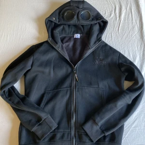 Svart hoodie med glasögon från C.P. Company - Svart hoodie från C.P. Company med dragkedja och ikoniska glasögon i huvan. Broderad logga på bröstet och två fickor framtill. Perfekt för dig som gillar streetwear och vill sticka ut.
