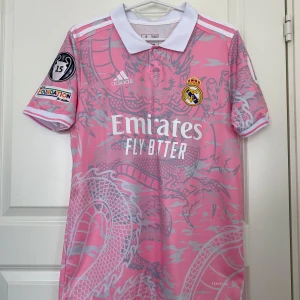Real Madrid rosa fotbollströja med drakmönster - Säljer en rosa Real Madrid fotbollströja med vitt drakmönster, klubbmärke och sponsortryck. Tröjan har vit krage, vita detaljer och Mbappé 9 på ryggen. Kortärmad modell med flera tryck på ärmarna.