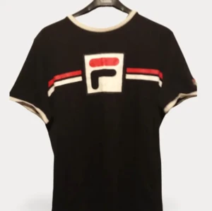 Svart t-shirt från Fila med logga - Svart t-shirt från Fila med stor logga framtill i vitt, rött och svart. T-shirten har vita och röda detaljer på bröstet och vita kanter vid ärmslut och halsringning. Perfekt för en sportig och avslappnad stil.