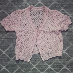 Ljusrosa spetskofta med kort ärm - Söt ljusrosa kofta med korta ärmar och vackert spetsmönster i geometriskt motiv. Perfekt att slänga över en topp för en extra touch. Koftan är öppen framtill och har vågiga kanter.