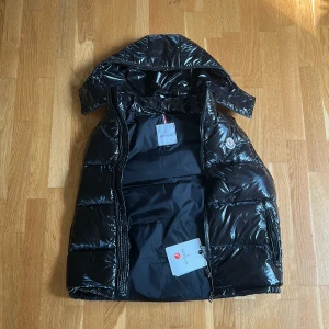 Svart dunvästfrån Moncler - Säljer en svart glansig dunväst från Moncler med huva och dragkedja. Jackan har en klassisk Moncler-logga på bröstet och är perfekt för kyligare dagar. Fylld med dun för extra värme.