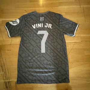 Real Madrid fotbollströja Vini Jr. 7 - Säljer en grå Real Madrid fotbollströja med Vini Jr. och nummer 7 på ryggen. Tröjan har korta ärmar, vita detaljer och klubbens emblem broderat på bröstet. Snyggt mönster över hela tröjan och klassisk passform.
