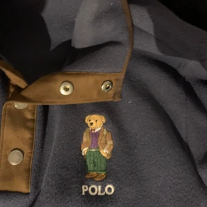 Blå fleecehoodie med björn från Ralph Lauren - Mörkblå fleecehoodie från Polo Ralph Lauren med broderad björn på bröstet. Jackan har tryckknappar i metall och detaljer i brunt tyg vid kragen. Perfekt för en avslappnad och stilren look.