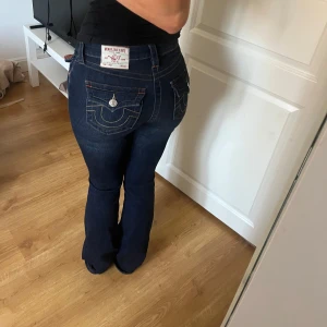 Mörkblå bootcut jeans från True Religion - Säljer ett par mörkblå jeans från True Religion, modell Becca Mid Rise Bootcut. Jeansen har snygga kontrastsömmar, dekorativa fickor med lock och silverfärgade knappar bak. Klassisk bootcut passform och normalhög midja. Använda väldigt fåtal ggr. Ord pris 1299kr, mitt pris 750kr