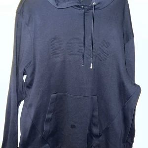Hugo BOSS - Säljer en mörkblå hoodie från BOSS med broderad logga på bröstet. Tröjan har huva med dragsko och en stor magficka framtill. Perfekt för en avslappnad stil. Samt en fläck som ni ser går säkert att få bort samt att snöret blivit lite paj storlek 3 xl passar xl