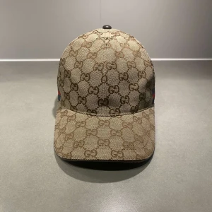 Beige GG keps - Snygg beige keps från Gucci med klassiskt GG-monogram över hela kepsen och en grön-röd rand på sidan. Justerbar rem baktill i skinn och insida i ljus textil. Perfekt för dig som gillar exklusiva accessoarer.