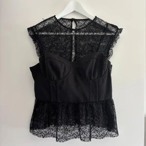 Svart spetsblus med korsett-detalj från Zara - Säljer en svart blus från Zara med transparent spets upptill och nedtill samt korsettliknande i mitten. Blusen har korta volangärmar. Toppen är helt ny och slutsåld på Zaras hemsida. Prislappen är kvar 