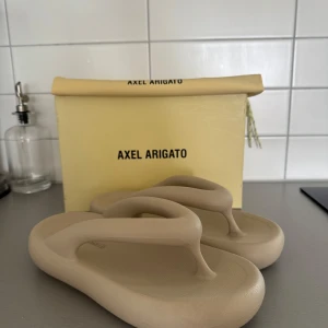 Beige sandaler från Axel Arigato - Chunky beige sandaler från Axel Arigato med modern, rundad form och tjock sula. Tillverkade i syntetmaterial och har en minimalistisk design med breda remmar. Perfekta för sommaren och ger en clean look till din outfit.