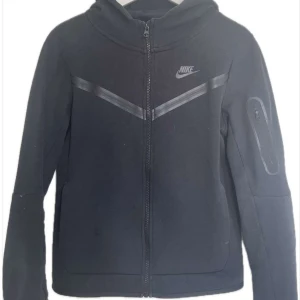 Svart hoodie från Nike - Svart hoodie från Nike med dragkedja framtill och diskret logga på bröstet. Jackan har en sportig look med svarta detaljer över bröstet och en praktisk ficka med dragkedja på ärmen. Perfekt för en aktiv vardag.