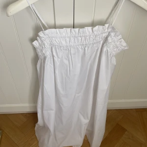 Vit axelbandslös klänning - Säljer en enkel och stilren vit axelbandslös klänning med resår upptill. Klänningen har smala axelband och en rak passform. Prislappen är kvar, då den aldrig kommit till användning!