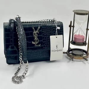 Svart axelväska från Yves Saint Laurent - Snygg svart axelväska från Yves Saint Laurent med krokodilpräglat mönster och silverfärgade detaljer. Väskan har en kedjerem och ikonisk YSL-logga framtill. Perfekt för att lyfta vilken outfit som helst.