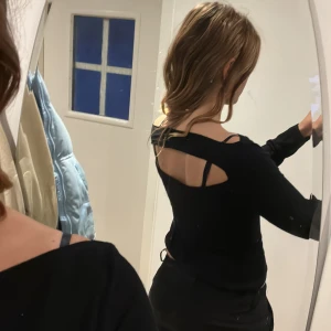 Svart croppad topp med öppen rygg och banddetalj - Säljer en svart långärmad croppad topp med unik öppen rygg och banddetalj. Toppen har en tight passform och är perfekt för dig som vill ha något stilrent men ändå med en twist.