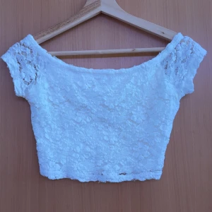 Vit spets croppad topp från Hollister - Säljer en söt vit croppad topp från Hollister i spets med blommigt mönster. Toppen har korta ärmar och en offshoulder-design som ger en romantisk känsla. Perfekt till sommaren!