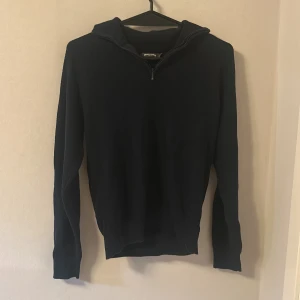 Mörkblå stickad tröja med dragkedja från Dressmann - Mörkblå stickad tröja från Dressmann i storlek S. Tröjan har lång ärm, hög krage och dragkedja vid halsen. Perfekt för lager-på-lager och enkel att matcha med olika outfits.