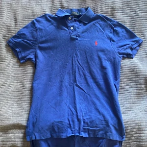 Blå pikétröja från Polo Ralph Lauren - Säljer en klassisk blå pikétröja från Polo Ralph Lauren med korta ärmar och krage. Tröjan har knappar vid halsen och den ikoniska röda loggan broderad på bröstet. 
