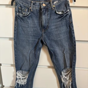 Blå jeans med slitningar - Säljer ett par blå jeans med slitna detaljer vid knäna och klassisk femficksdesign. Jeansen har normal passform och är tillverkade i denim. Perfekta för en avslappnad stil.