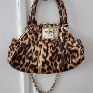 Leopardmönstrad handväska från Guess - Säljer en snygg leopardmönstrad handväska från Guess med gulddetaljer och kedjehandtag. Väskan har ett stort metallmärke framtill och innerfoder med mönster samt dragkedjefack. Perfekt för dig som vill sticka ut med din stil.