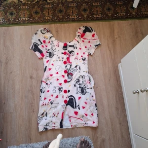 Vit kortklänning med hjärtan och tecknat mönster - Säljer en söt vit kortklänning med röda hjärtan och svart tecknat mönster. Klänningen har puffärmar, stora röda knappar framtill och en rundad urringning. Perfekt för dig som gillar unika och lekfulla plagg.