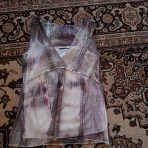 Lila transparent ärmlös topp med paljettdetalj - Säljer en lila transparent ärmlös topp med v-ringning och dekorativa paljetter framtill. Toppen har ett skirt tyg och subtila ränder i olika nyanser av lila och vitt. Perfekt för att skapa en unik och trendig look.