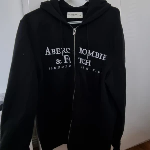 Svart hoodie med dragkedja från Abercrombie & Fitch - Svart hoodie från Abercrombie & Fitch med vit logga på bröstet. Tröjan har huva, dragkedja framtill och är tillverkad i mjuk fleece. Perfekt för en avslappnad stil.