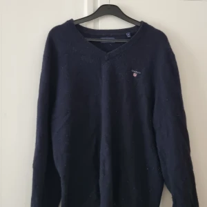 Mörkblå v-ringad tröja från GANT - Mörkblå v-ringad tröja från GANT med diskret broderad logga på bröstet. Klassisk modell med långa ärmar och ribbade muddar. Perfekt för en stilren och enkel look.