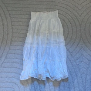 Vit långkjol från Indiska - Säljer en vit långkjol från Indiska med smock i midjan och flera volanger. Kjolen är luftig och har ett romantiskt uttryck med fina detaljer. Perfekt för varma dagar och enkel att matcha med olika toppar.