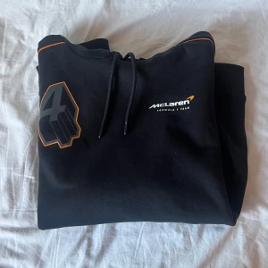 Svart hoodie från McLaren Racing LN4 - Säljer en svart hoodie från McLaren Formula 1 Team med LN4-tryck och Lando Norris-detaljer. Tröjan har huva , tryck på både bröst och rygg samt orange detaljer. Perfekt för racingfans!
