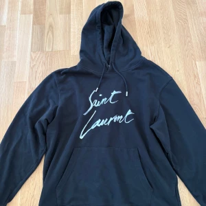 Svart hoodie från Saint Laurent - Säljer en svart hoodie från Saint Laurent med vit logga framtill. Klassisk modell med huva och känguruficka. Perfekt för en stilren och avslappnad look. Storlek XL men sitter som L.