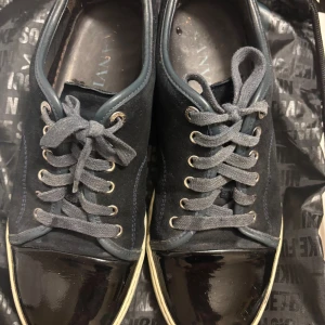 Svarta sneakers från Lanvin med lackdetalj - Säljer ett par svarta sneakers från Lanvin med snygga lackade tår och grå snörning. Skorna har en platt vit sula och klassisk rund tå. Perfekta för dig som gillar stilrena och exklusiva sneakers. Dom är i 8/10 skick inga speciella skador bara lite repor på glasen.