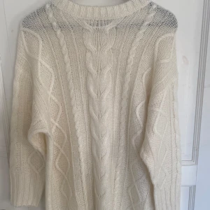 Vit stickad kabelstickad klänning - Säljer en mysig vit stickad klänning med klassiskt kabelstickat mönster och rund hals. Perfekt för kyliga dagar och enkel att styla till olika outfits. Använd en gång.