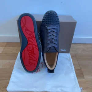 Svarta sneakers med nitar från Christian Louboutin - Säljer ett par gråa sneakers från Christian Louboutin med ikoniska röda sulor och gråa nitar på tån. Skorna har snörning och är tillverkade i skinn med en rund tå. Perfekta för dig som vill sticka ut med en lyxig och edgy stil.