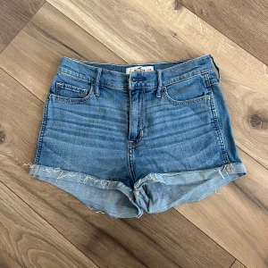 Jeansshorts från Hollister - Blå jeansshorts från Hollister med klassisk femficksdesign och råa, fransiga benslut. Shortsen har uppvikta kanter. Ikoniska bakfickor med dekorativa sömmar. Välanvända.