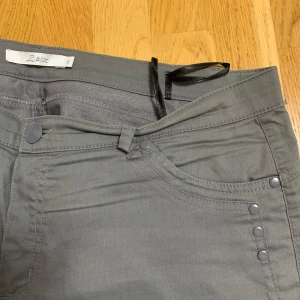 Gråa halvshorts från Zizzi - Snygga gråa halvshorts från Zizzi med klassisk femficksdesign och silverfärgade nitar. Shortsen har normal passform och är tillverkade i ett mjukt bomullsmaterial med lite stretch. Perfekta för en stilren vardagslook.