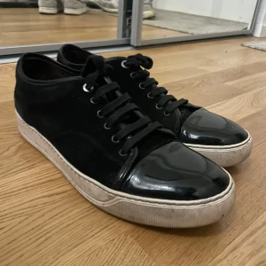 Svarta sneakers med lackad tå och snörning - Säljer ett par svarta sneakers med snygg lackad tå och snörning. Skorna har en vit platt sula och är tillverkade i en kombination av mocka och lackmaterial. Perfekta för dig som gillar stilrena och moderna skor.