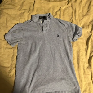 Ljusgrå pikétröja från Polo Ralph Lauren - Säljer en klassisk ljusgrå pikétröja från Polo Ralph Lauren med korta ärmar och krage. Tröjan har knappar vid halsen och den ikoniska broderade loggan på bröstet. Perfekt för en stilren och avslappnad look.