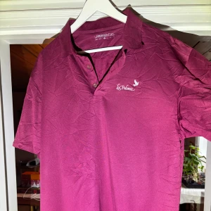 Vinröd pikétröja från Nike Golf - Säljer en vinröd pikétröja från Nike Golf med Dri-FIT-material. Tröjan har korta ärmar, krage och knappar framtill. Broderad Nike-logga på ärmen och La Paloma-tryck på bröstet. Perfekt för golf eller vardag.