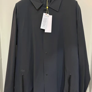 Svart väst med huva från Lacoste - Säljer en svart väst från Lacoste med huva och dragkedja framtill. Västen har två fickor med dragkedja och en stilren design utan ärmar. Perfekt för lager-på-lager och enkel att matcha med olika outfits. En kappa kommer med västen