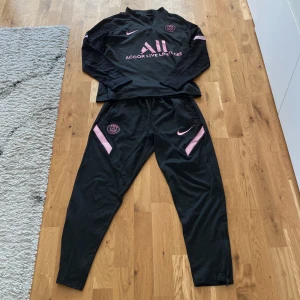 Svart PSG fotbollsträningsoverall från Nike - Säljer en svart långärmad PSG på byxorna är det ingen dragkedja. träningsoverall från Nike med rosa detaljer och loggor. Tröjan har halv dragkedja och byxorna har resår i midjan. Materialet är lätt och andas, perfekt för träning eller uppvärmning.