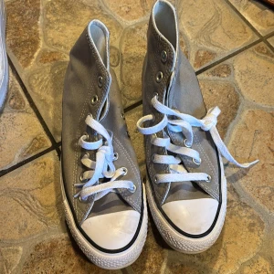 Grå höga Converse All Star sneakers - Säljer ett par klassiska grå höga Converse All Star med vit tå och vita snören. Skorna har den ikoniska loggan på sidan och svart rand runt sulan. Perfekta för dig som gillar tidlös streetstil.