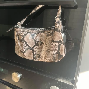 Axelväska i ormmönstrad syntet - Säljer en snygg axelväska med ormmönster i beige och bruna toner. Väskan har dragkedja upptill och en kort axelrem. Perfekt för att lyfta vilken outfit som helst med lite edge.