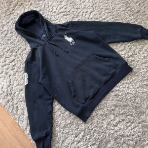 Mörkblå hoodie från Polo Ralph Lauren - Säljer en mörkblå hoodie från Polo Ralph Lauren med stor vit logga på bröstet och broderad siffra på ärmen. Tröjan har huva med dragsko och en stor magficka framtill. Perfekt för en avslappnad stil. OBS finns 2 fläckar, skickar privat bilder. Mått: längd: 59cm armhåla till armhåla 53cm