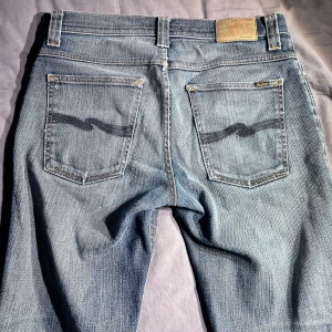 Ljusblå jeans från Nudie Jeans - Säljer ett par klassiska ljusblå jeans från Nudie Jeans med raka ben och snygga kontrastsömmar. Jeansen har fem fickor och subtila slitningar på bakfickorna. Perfekta för dig som gillar en tidlös denimlook.
