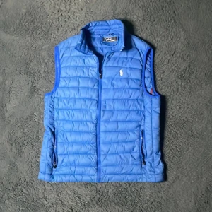 Blå dunväst från Polo Ralph Lauren - Snygg blå dunväst från Polo Ralph Lauren med broderad logga på bröstet. Västen har dragkedja framtill och två fickor med dragkedja. Perfekt för lager-på-lager och håller dig varm under kyliga dagar.