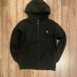 Polo Ralph Lauren Zip Hoodie – Svart – Strl M - Säljer en snygg och stilren Polo Ralph Lauren zip hoodie i storlek M. Färg: Svart med vit broderad logga på bröstet. 💯 Original.  ✅ Skick: Mycket bra – sparsamt använd och helt fri från fläckar, hål eller tvättpåverkan. 📏 Normal passform, perfekt till både vardag och streetwear-stil. 🧵 Mjuk bomullsblandning, med dragkedja och kängurufickor.  ⸻   💬 Hör gärna av dig vid frågor! 
